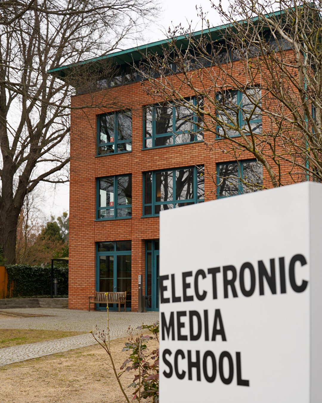 Im Vordergrund weißes Schild mit dem Schriftzug "Electronic Media School", dahinter das ems-Schulgebäude aus Backstein mit grünen Fenstern.