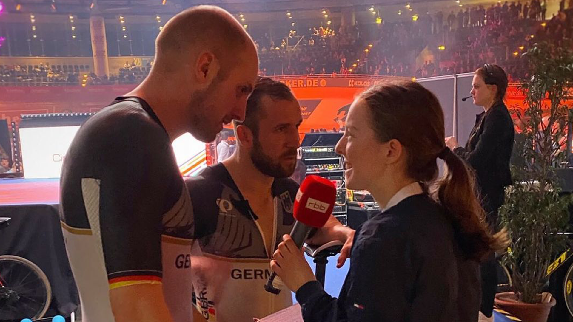 Junge Frau mit rbb-Mikrofon interviewt beim Berliner Sechstagerennen zwei Radsportler.